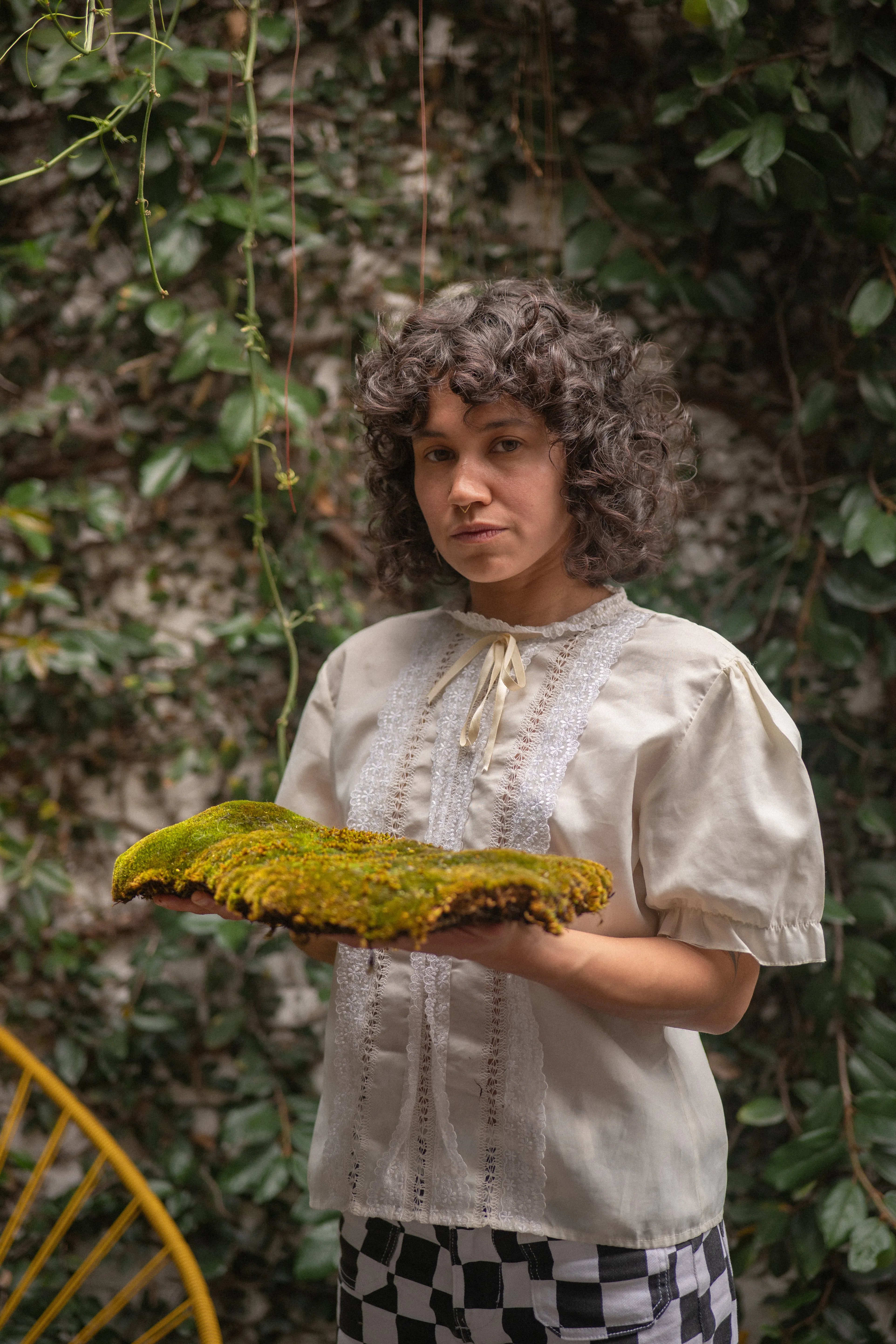 dark curly-haired woman with a septum piercing holding moss on a terrace in mexico city, intense gaze
