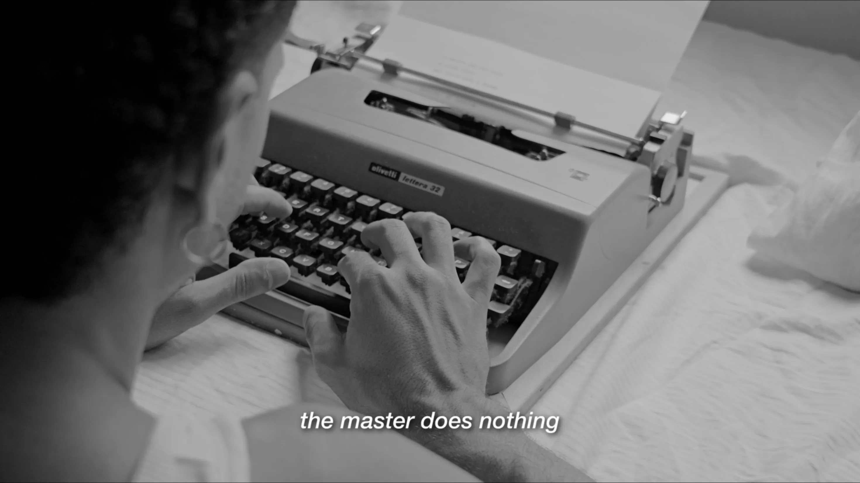 black and white, over the shoulder, a man typing on an olivetti typewriter on a bed, subtitle: the master does nothing