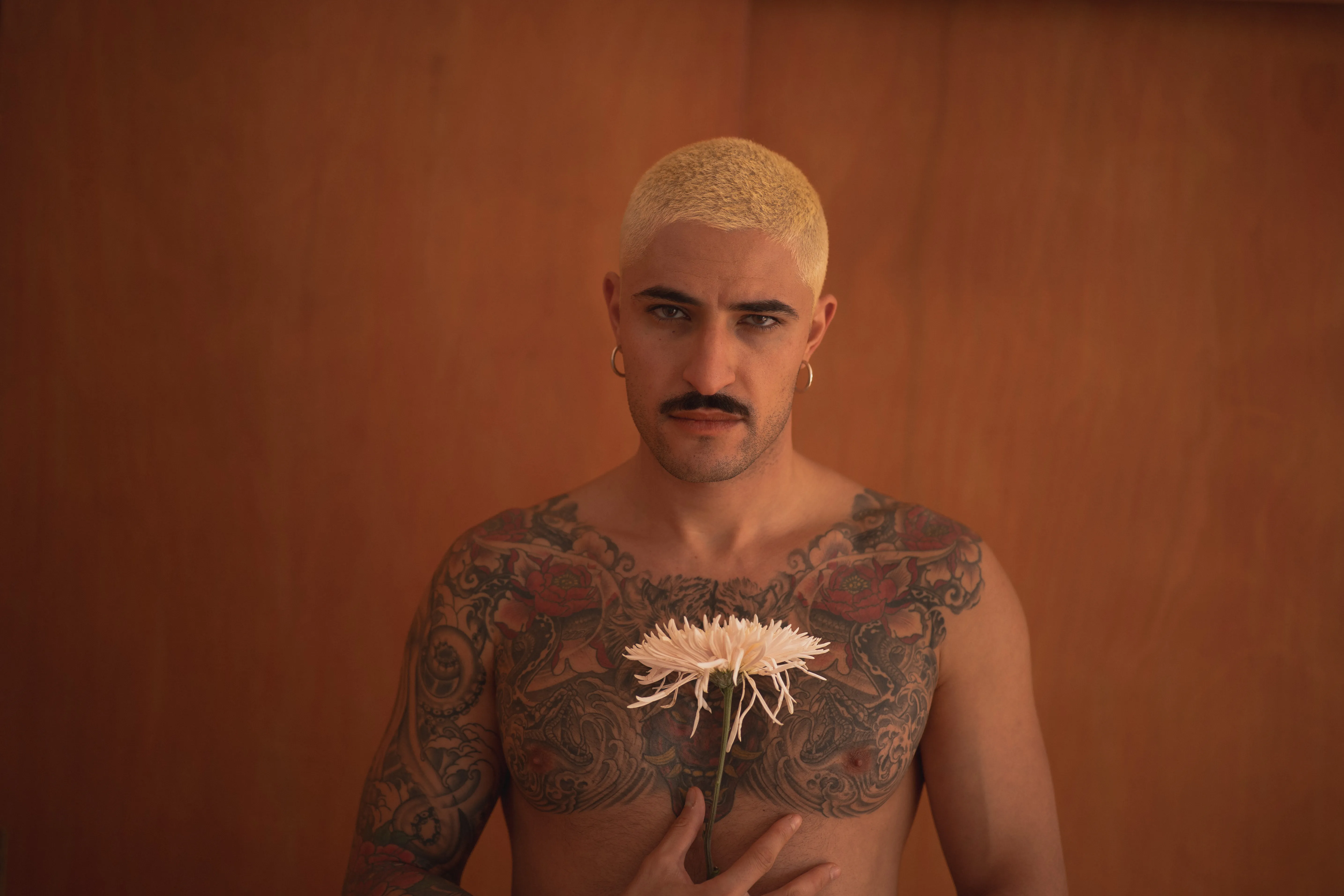 tattooed boy with short bleached hair holding a spider chrysanthemum against an orange wall, peony tattoos, intense eyes, moustache and earrings