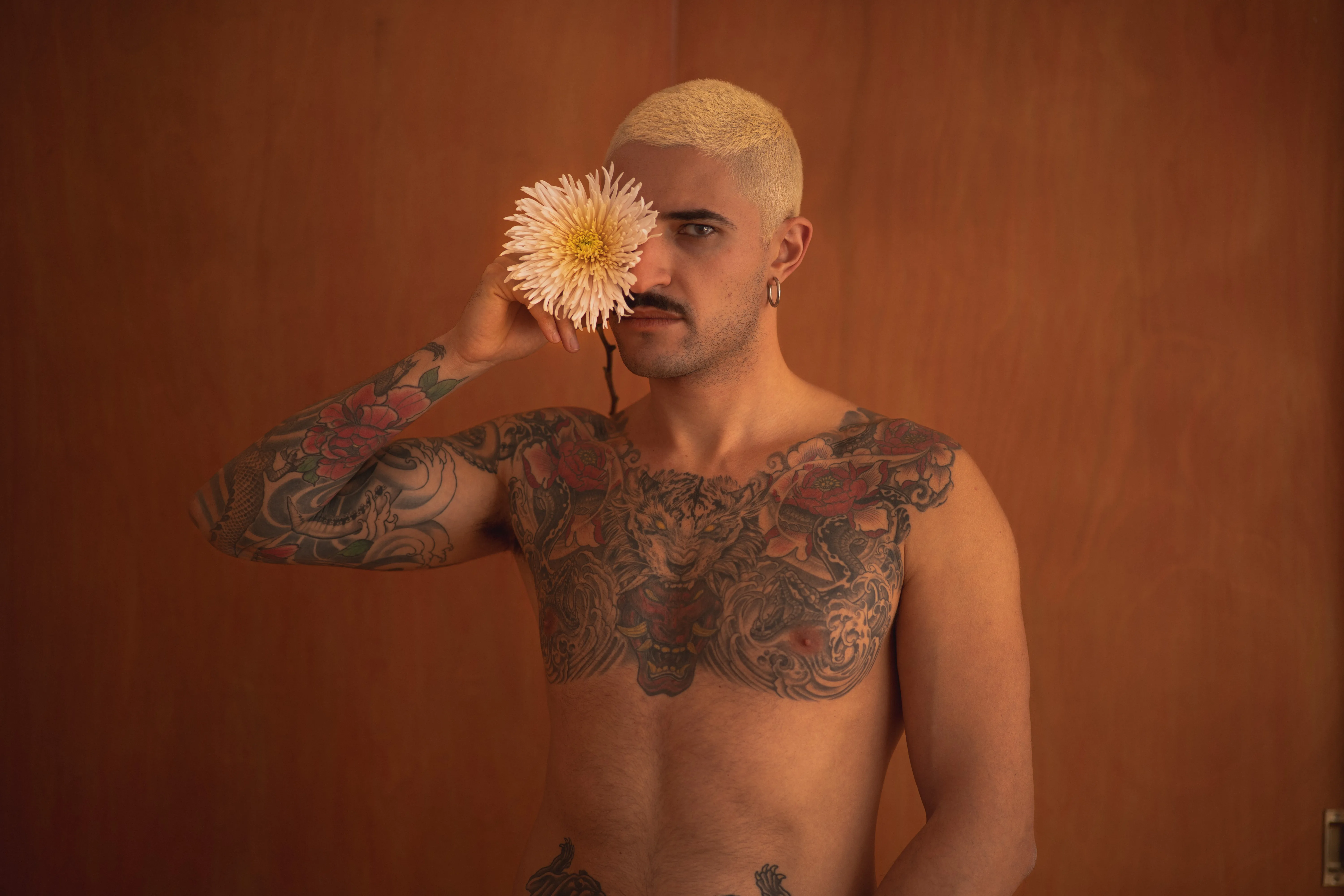 tattooed boy with short bleached hair holding a spider chrysanthemum against an orange wall, peony tattoos, intense eyes, moustache and earrings (2)