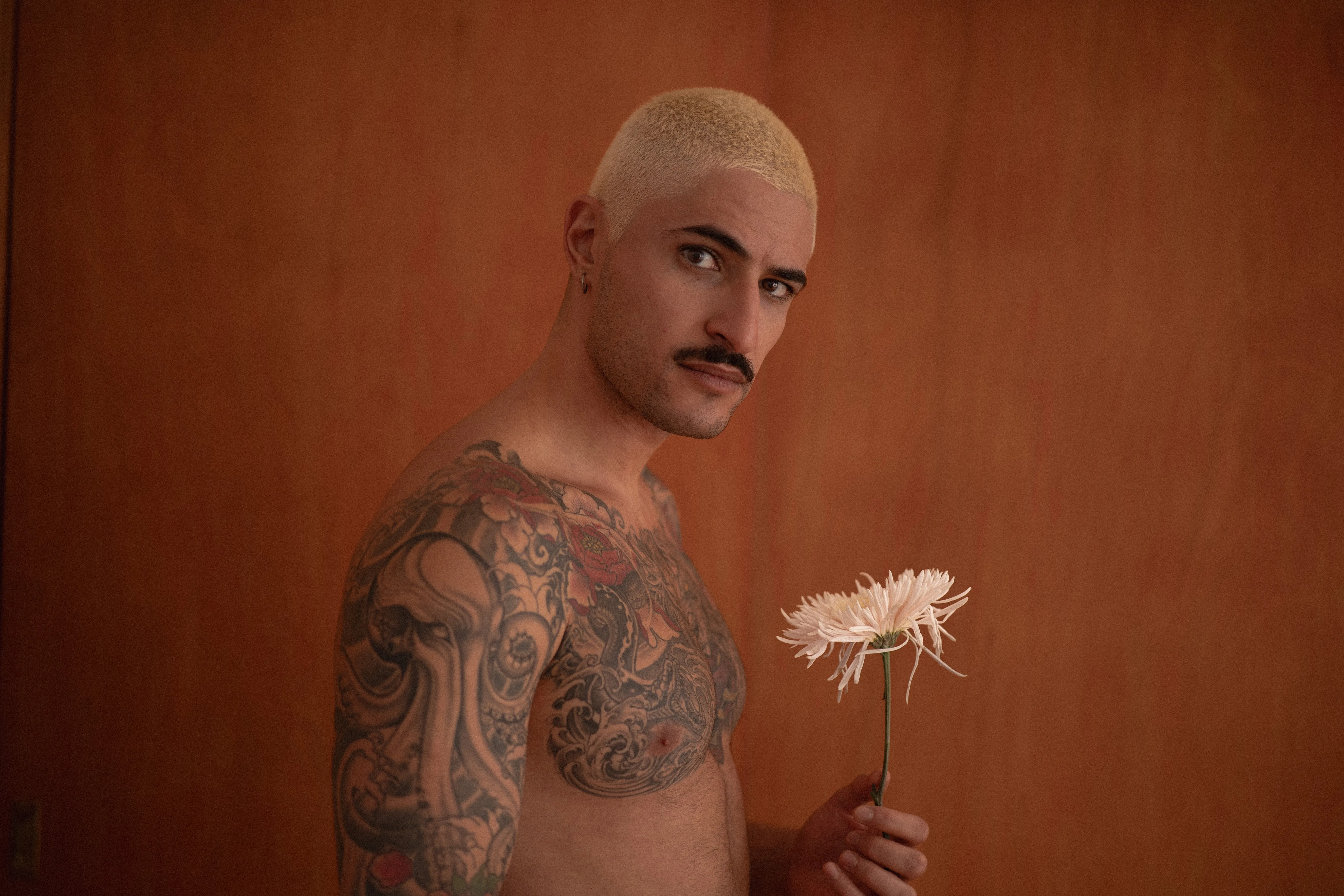 tattooed boy with short bleached hair holding a spider chrysanthemum against an orange wall, peony tattoos, intense eyes, moustache and earrings (3)