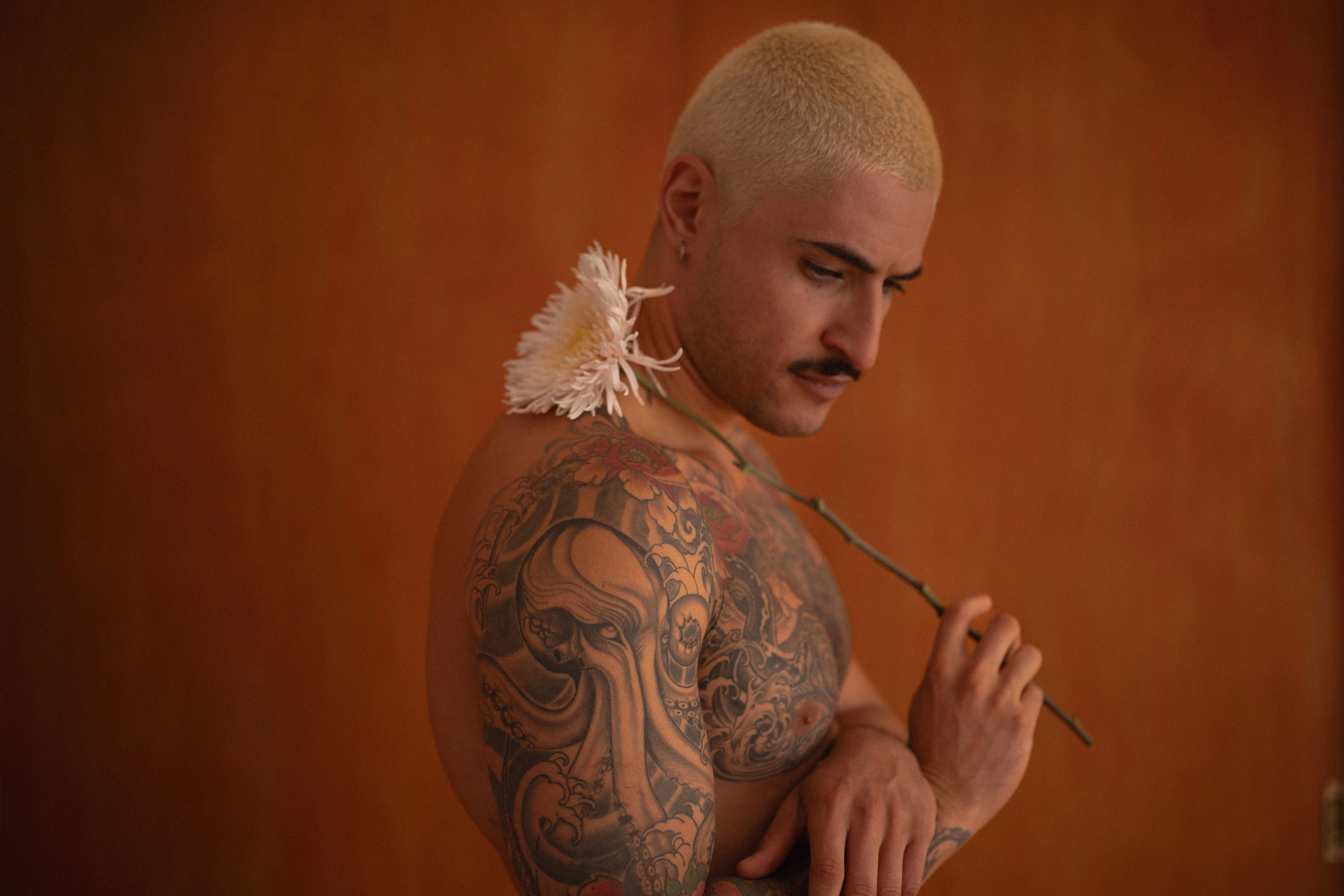 tattooed boy with short bleached hair holding a spider chrysanthemum against an orange wall, peony tattoos, intense eyes, moustache and earrings (4)