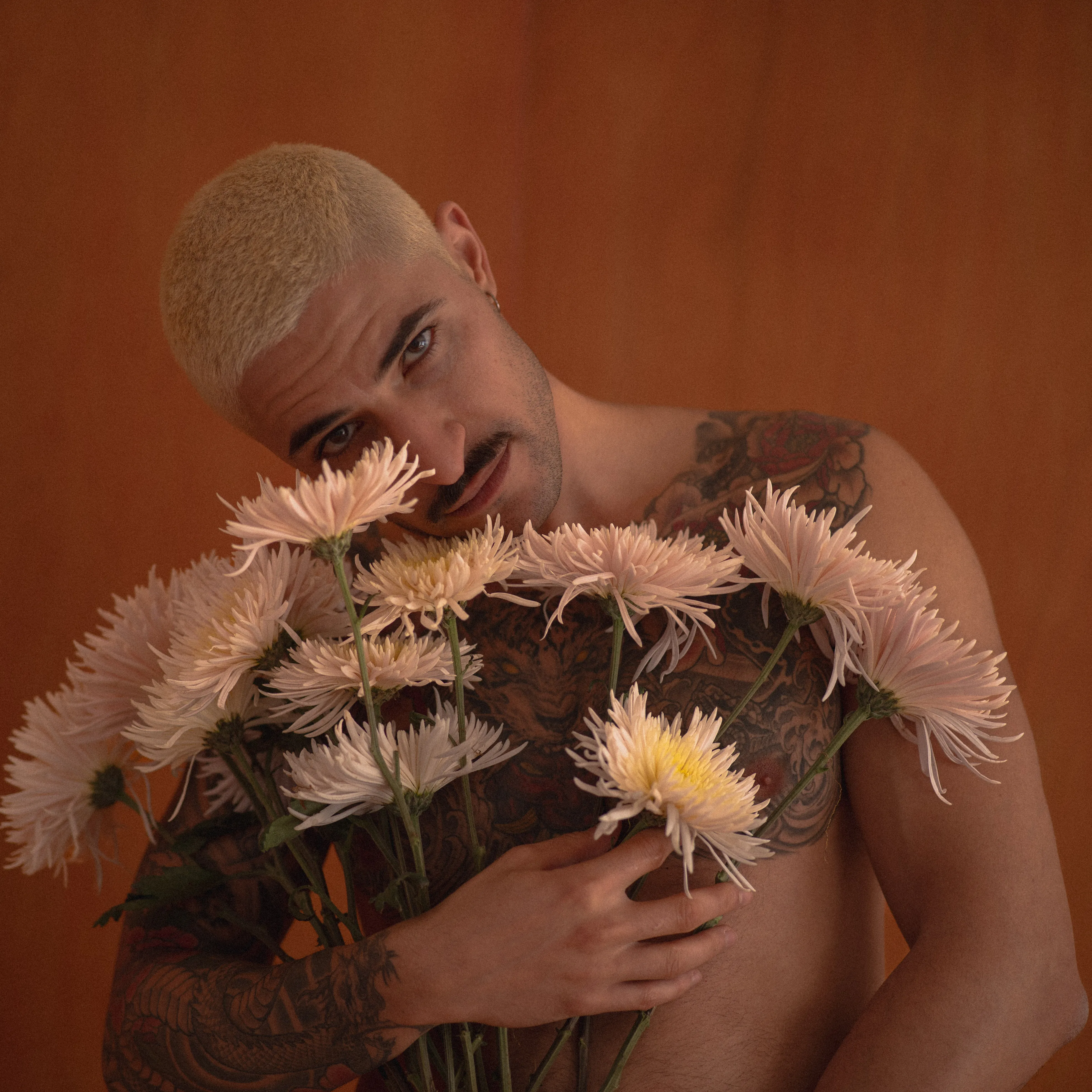 tattooed boy with short bleached hair holding a spider chrysanthemum against an orange wall, peony tattoos, intense eyes, moustache and earrings (5)